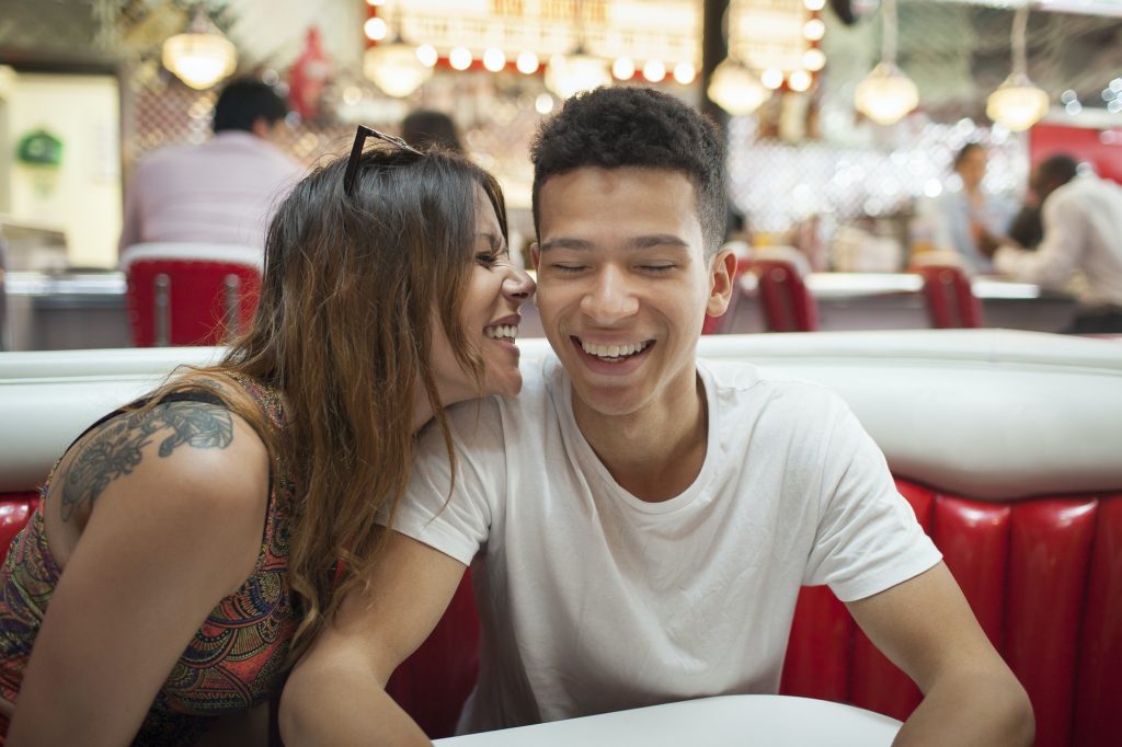 Young couple sitting in diner, young woman whispering in man's ear, laughing